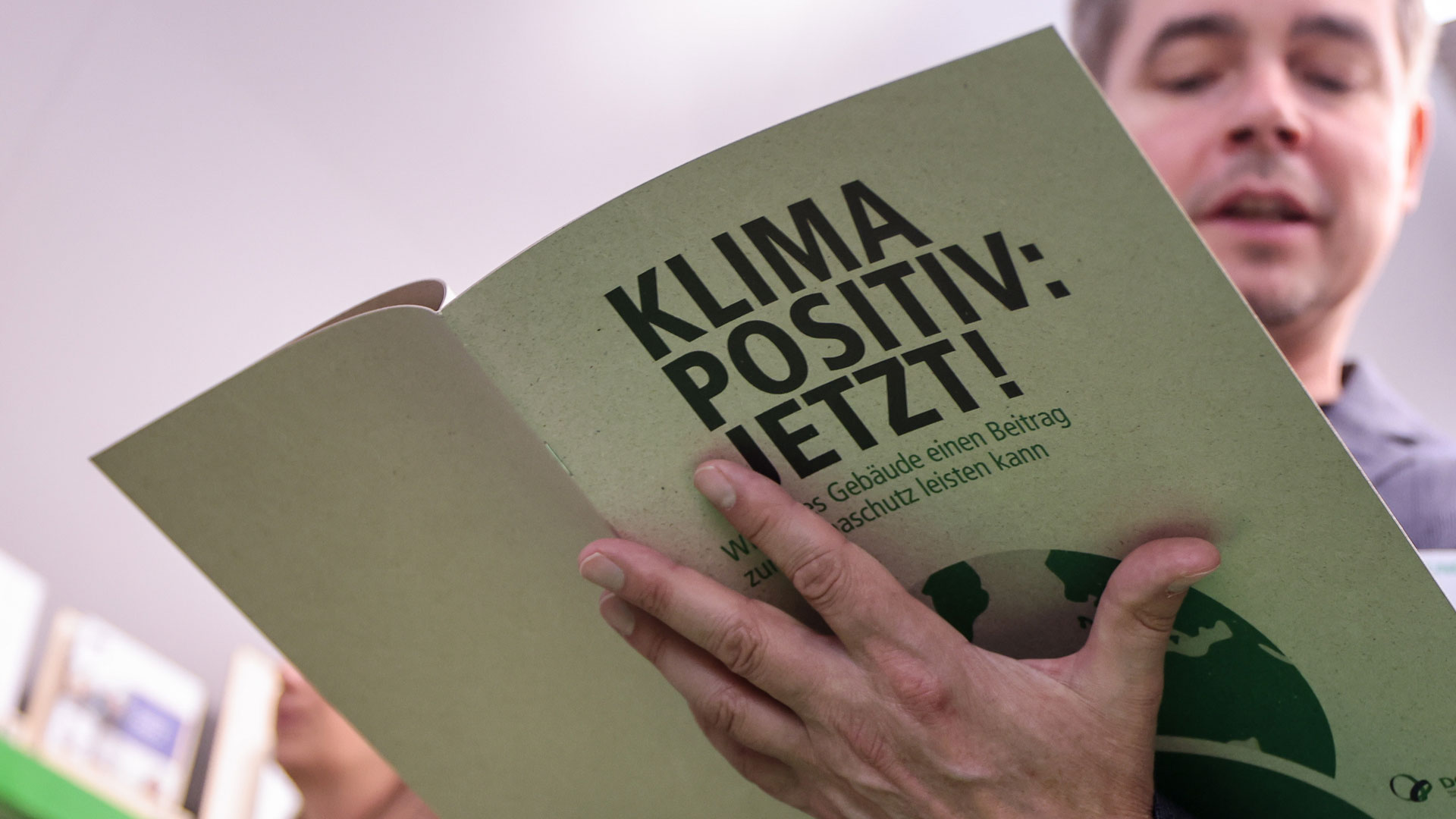 Das Bild zeigt ein Foto einer aufgeklappten Borschüre. Auf dem Titel steht "Klimapositiv: Jetzt!". Die Borschüre liegt aufgeklappt in der Hand einer Person, deren Gesicht im Hintegrund verschwommen zu sehen ist.