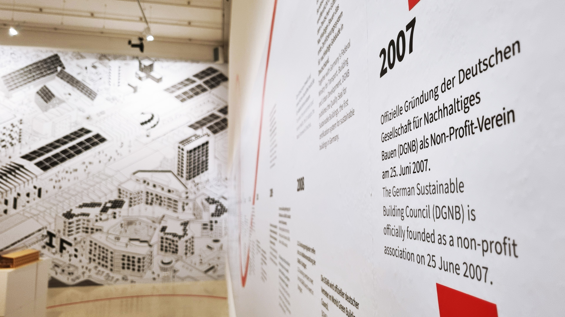 Das Bild zeigt einen Ausschnitt der DGNB Ausstellung bei Aedes. Zu sehen ist auf der rechten seite ein angeschnittener Zeitstrahl. Der Blick in den Raum gibt die Sicht auf ein großes schwarz-weißes Wimmelbild frei.