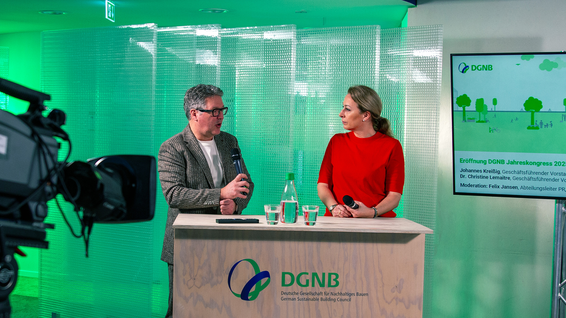 Mann und Frau stehen hinter einem Podium, von links ist eine Kamera auf sie gerichtet, im Hintergrund erscheint eine Begrüßungsfolie zum DGNB Jahreskongress 2025. Hintergrund ist grün ausgeleuchtet.