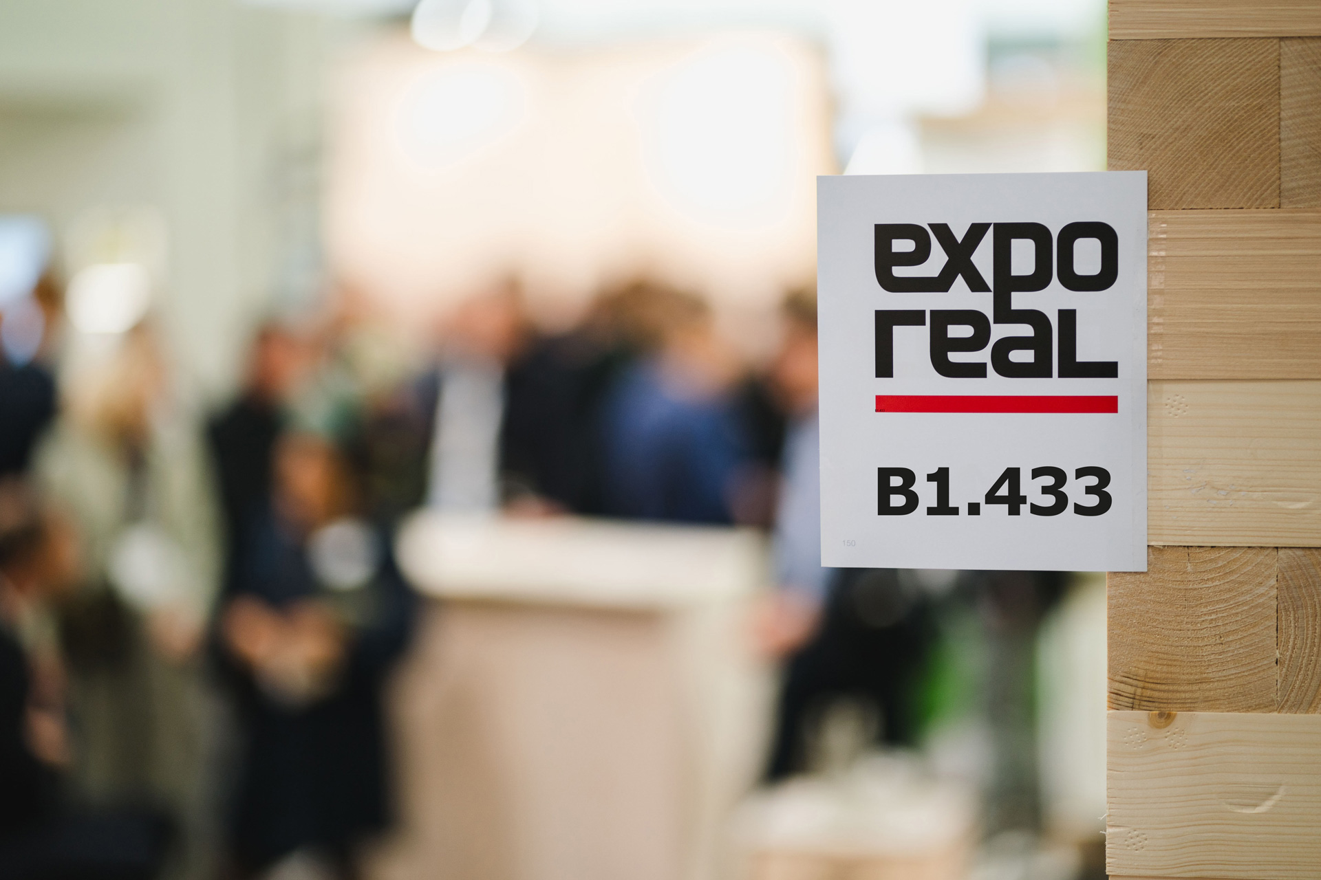 Expo Real Das Foto zeigt ein Wimpel, das an einer Holzwand klebt. Auf dem weißen Wimpel ist das Logo der Expo Real abgebildet sowie die Standnummer B1.433. Im Hintergrund ist vorschwommen ein besuchter Messestand zu sehen.