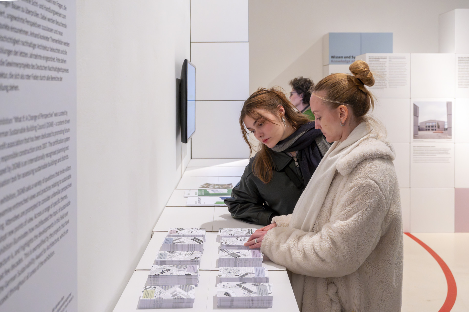 Zwei Personen, gekleidet in Wintermänteln, stehen in den Räumlichkeiten der DGNB Ausstellung bei Aedes und betrachten die ausgelegten Postkarten zur Ausstellung.