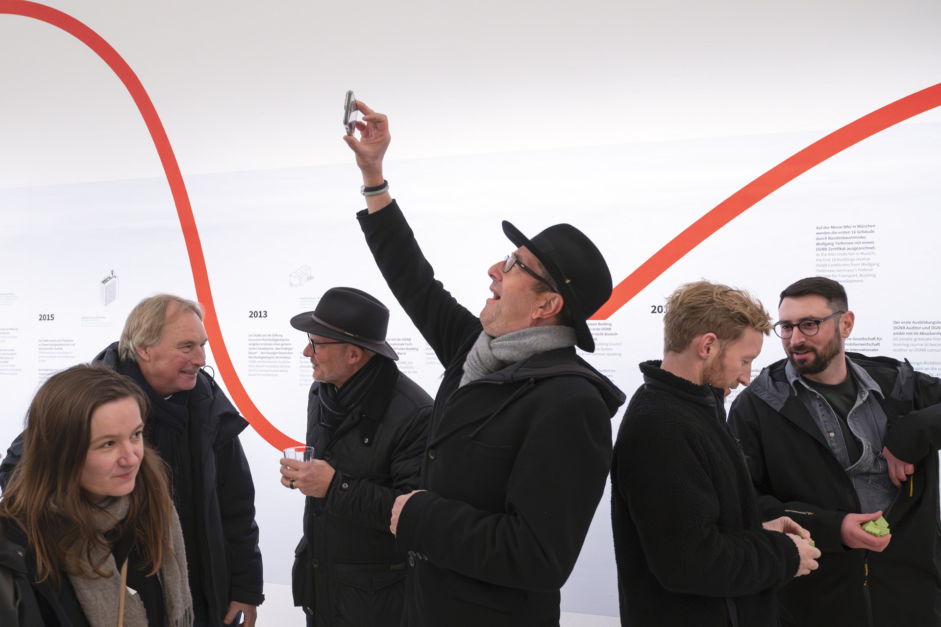 Impressionen von der Eröffnung der DGNB Ausstellung bei Aedes. Sechs Personen in winterlicher Kleidung sind vor dem großen Zeitstrahl der Ausstellung an der Wand fotografiert. Manche unterhalten sind. Einer hat sein Smartphone hochgehoben.