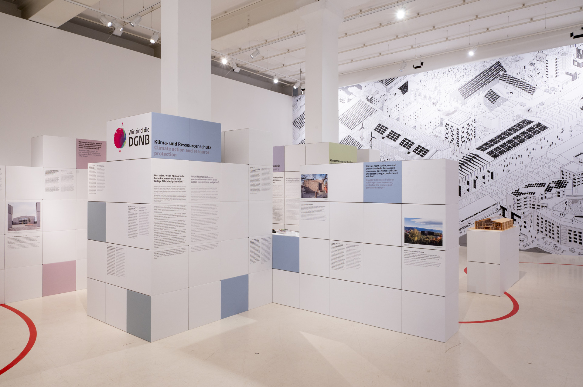 Einblick in die DGNB Ausstellung bei Aedes. Zu sehen sind Ausstellungswände aus Pappwürfeln sowie eine geschwungene rote Linie. die sich durch den weißen Raum zieht. Im Hintergrund ist an der Wand das große Wimmelbild zu erkennen.
